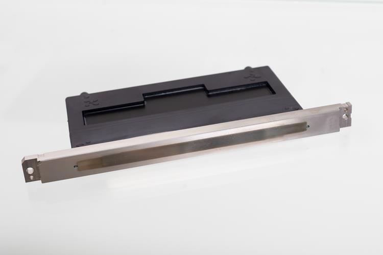 Photograph of a black and silver electronic component, likely a card reader or slot interface, placed on a white surface. The component features a rectangular black plastic housing with a horizontal slot and a silver metallic front panel with mounting holes on each end.