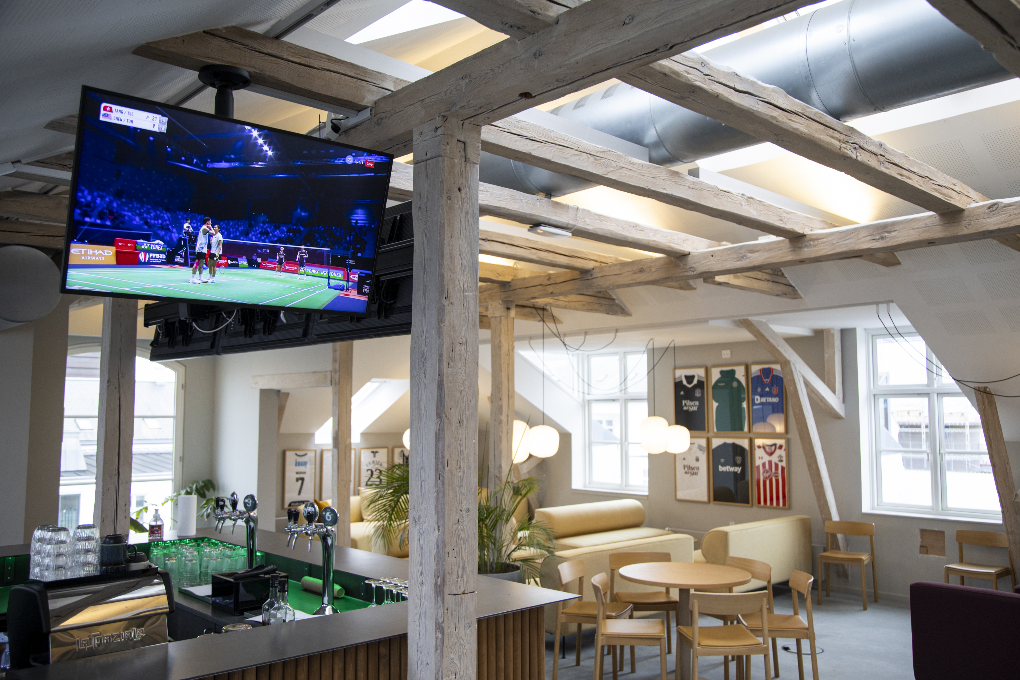 Lyst loungeområde med bar, tv‑skærme og afslappede siddepladser under synlige loftsbjælker.