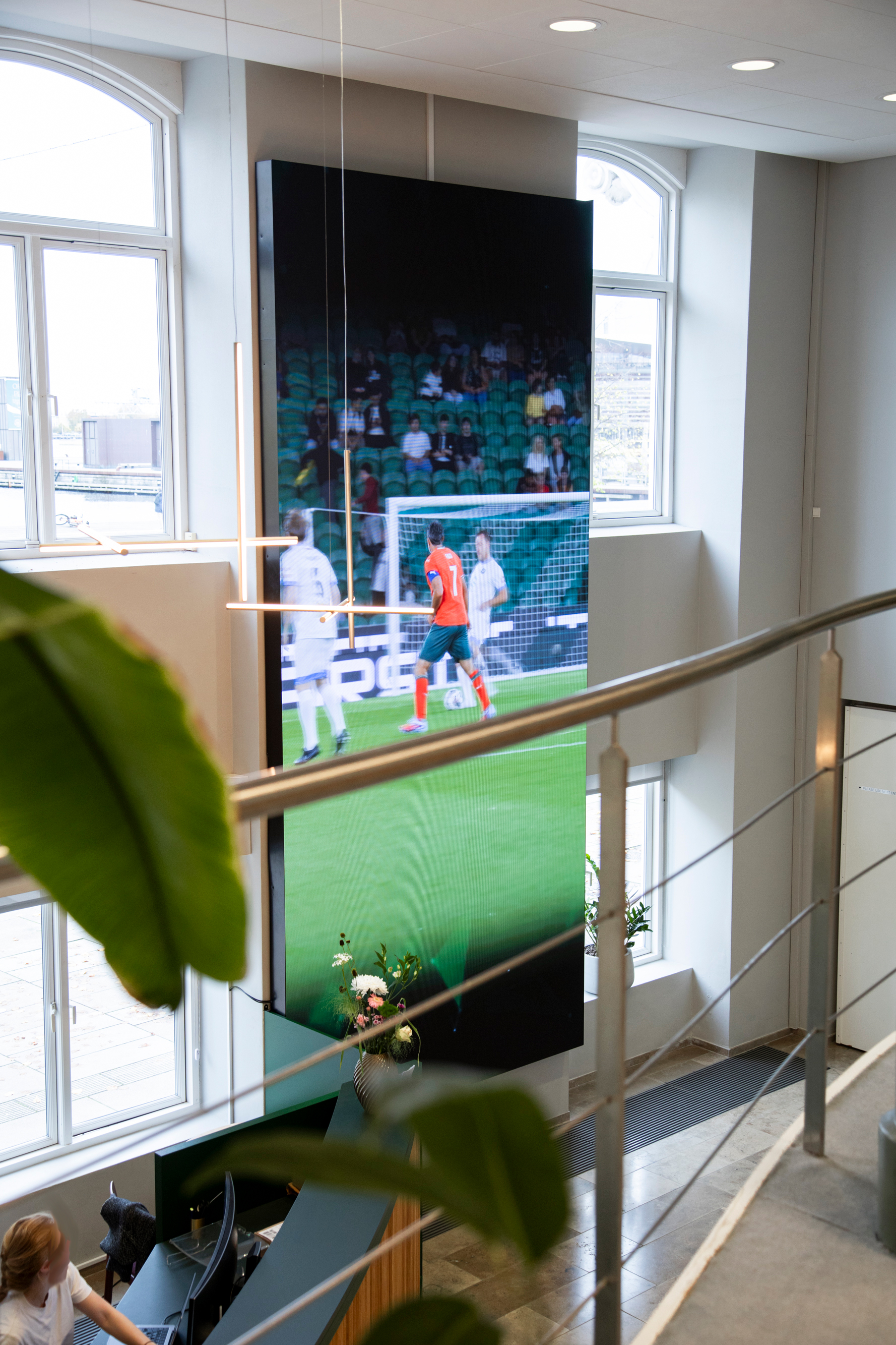 Upper‑floor view of a modern office interior featuring a tall digital screen and large windows.